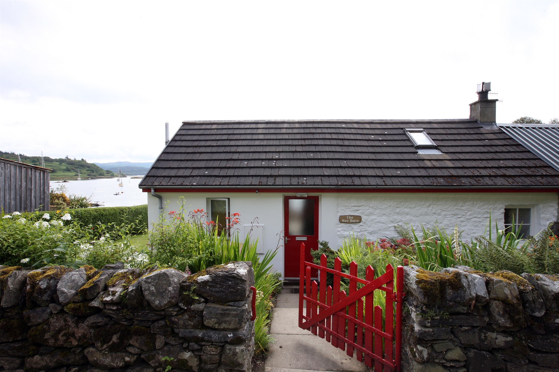 The Wee Barn's whitewashed exterior with red gate and stone wall, bay in the distance
